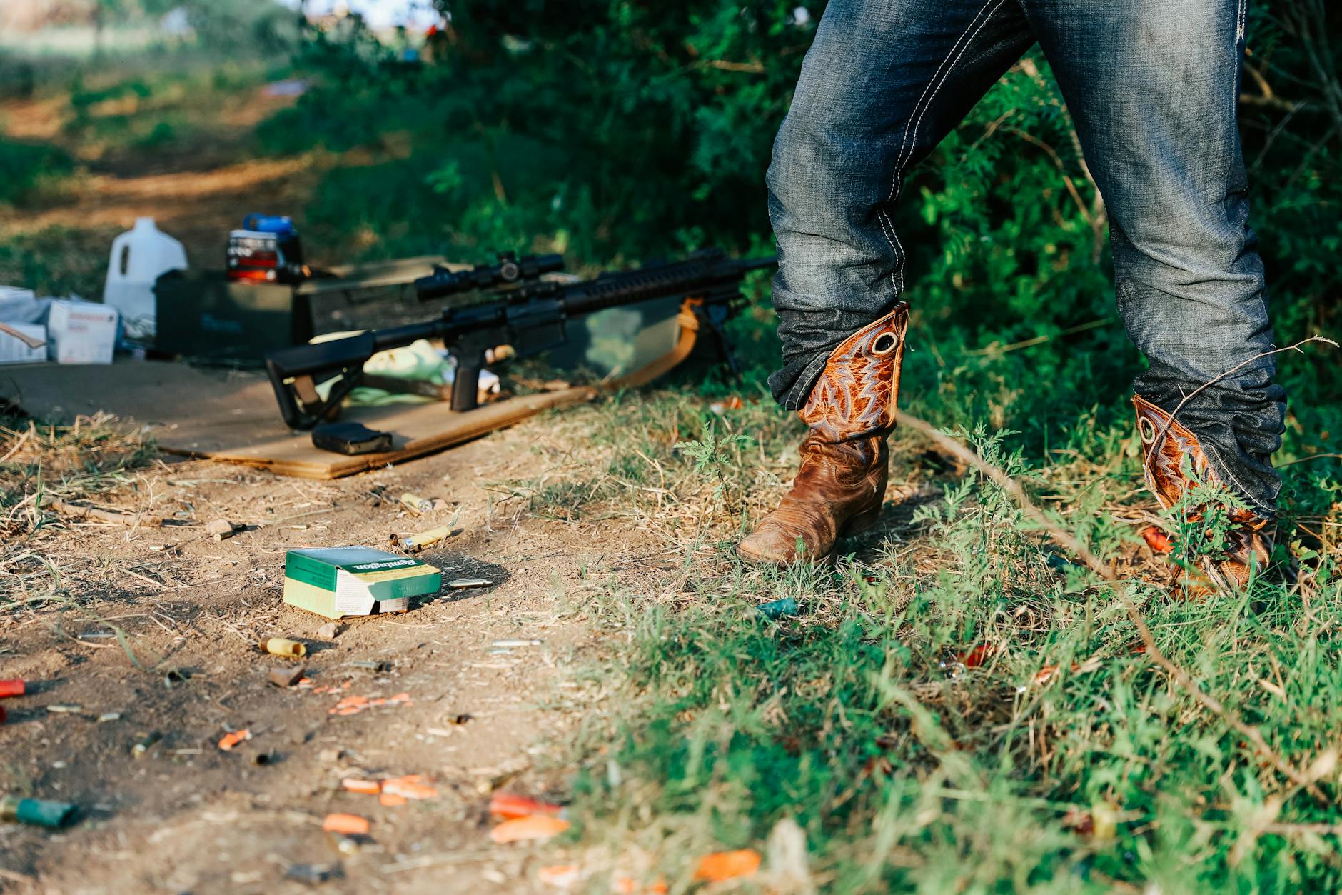 A person wearing cowboy boots stands outdoors, rifle on the ground surrounded by ammunition.